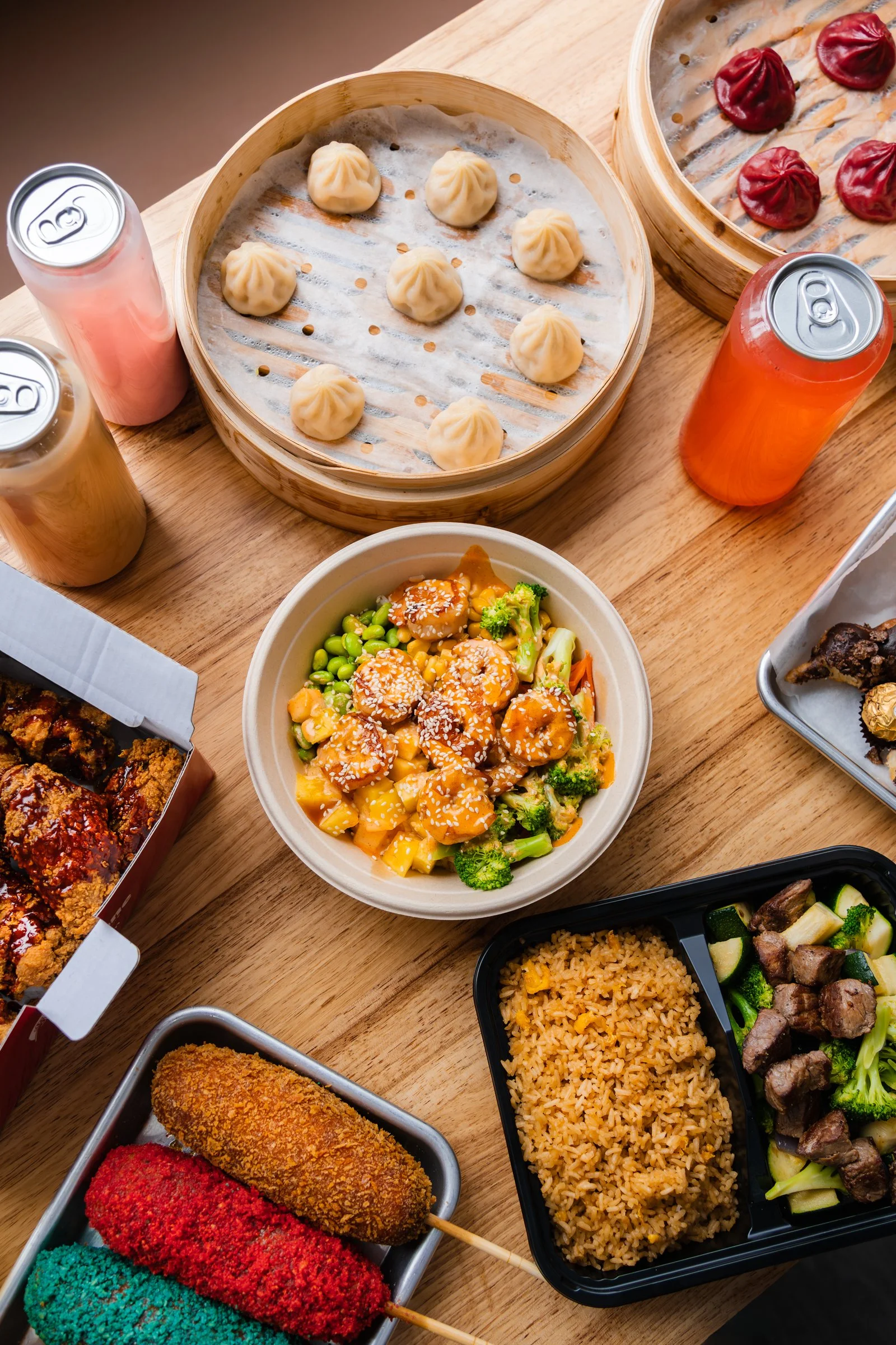 A variety of Asian dishes including steamed dumplings in bamboo steamers, fried chicken, colorful skewers, salad, rice, and stir-fried vegetables on a wooden table
